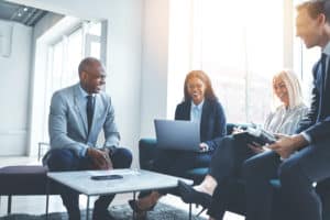 Diverse group of smiling businesspeople talking together while sitting on a sofa in a bright modern office